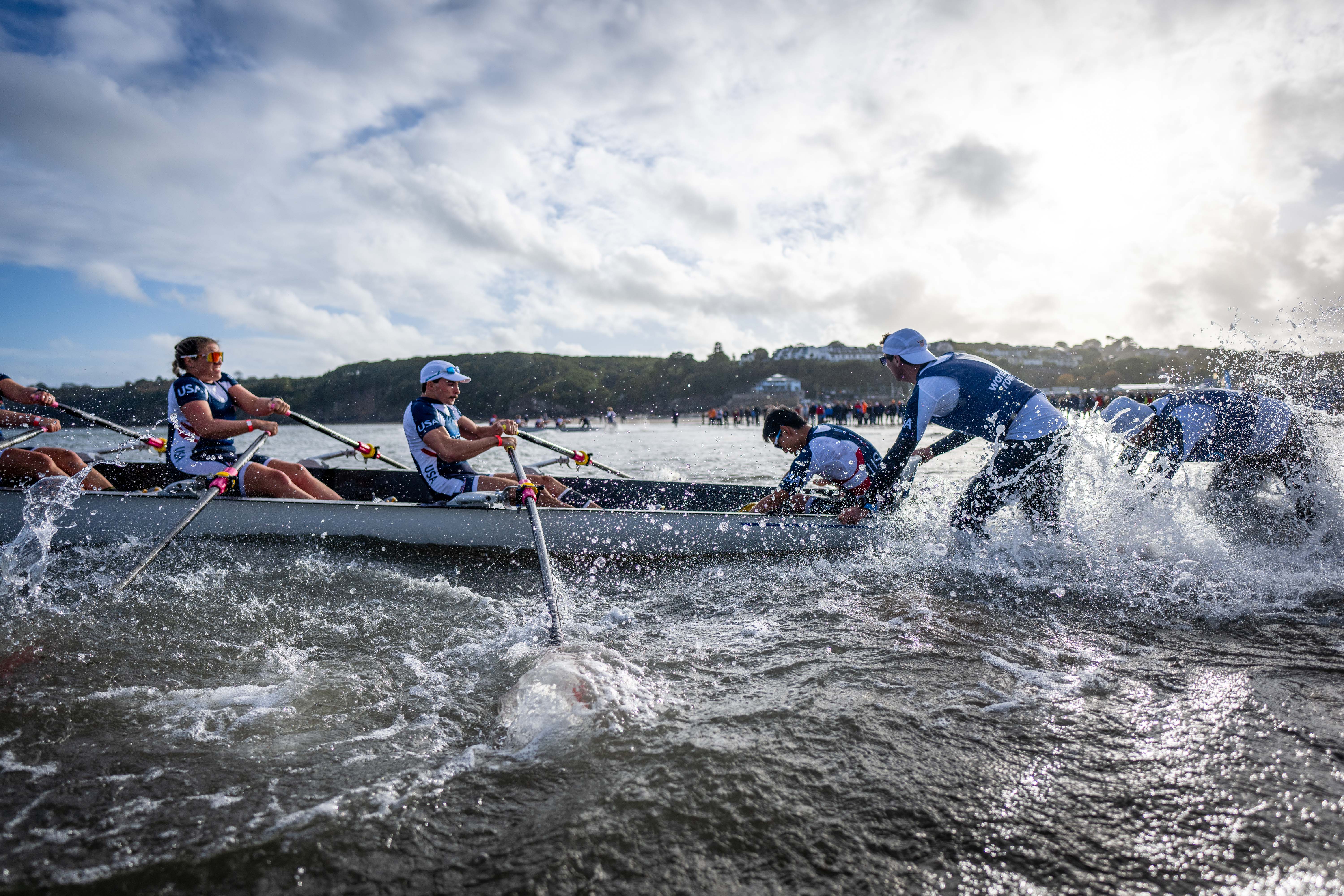 Two rowers in a boat set off into the water, with a group of people pushing them out into the water.