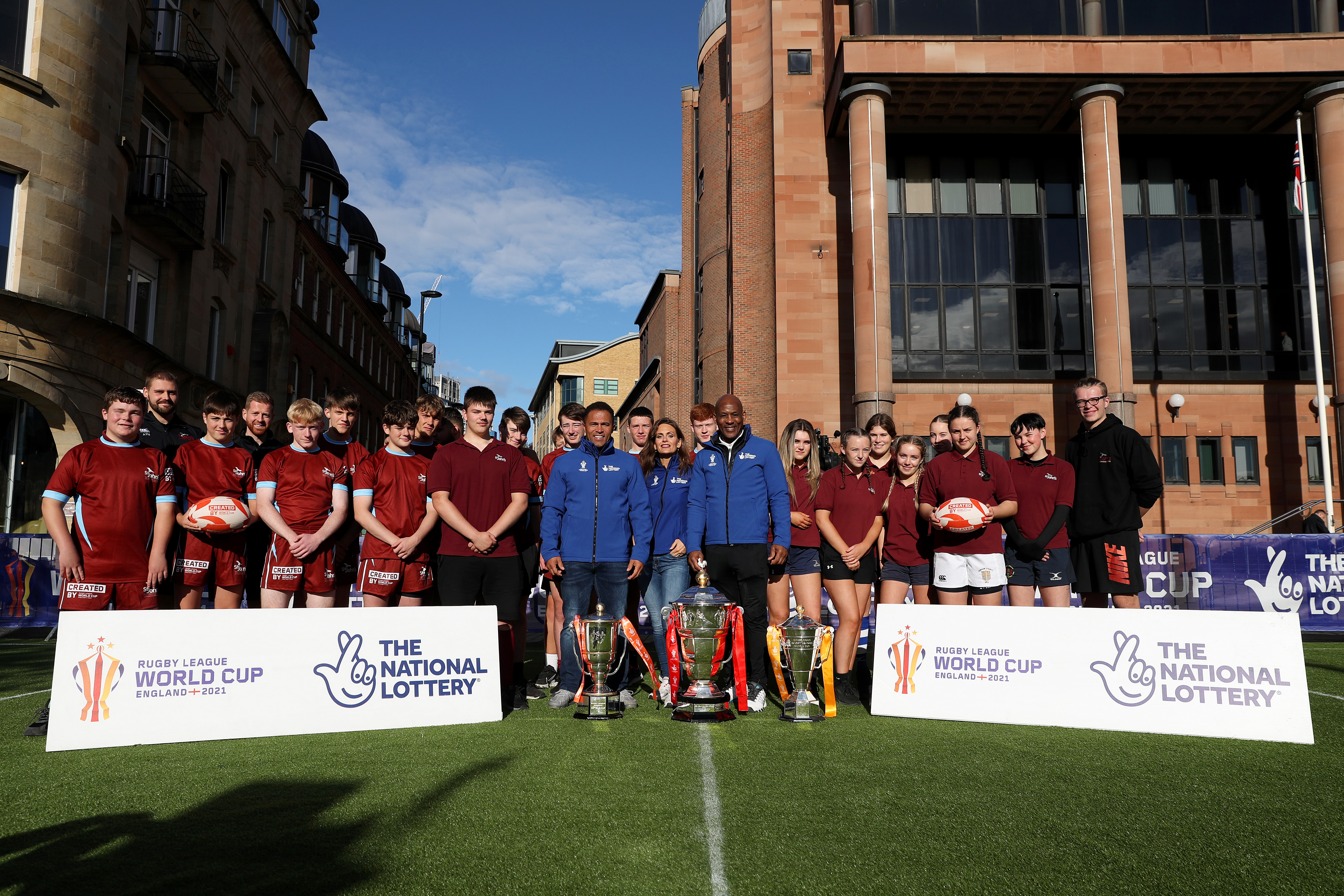 A group of people stand in front of a sign with the Rugby League World Cup logo and The National Lottery logo on.