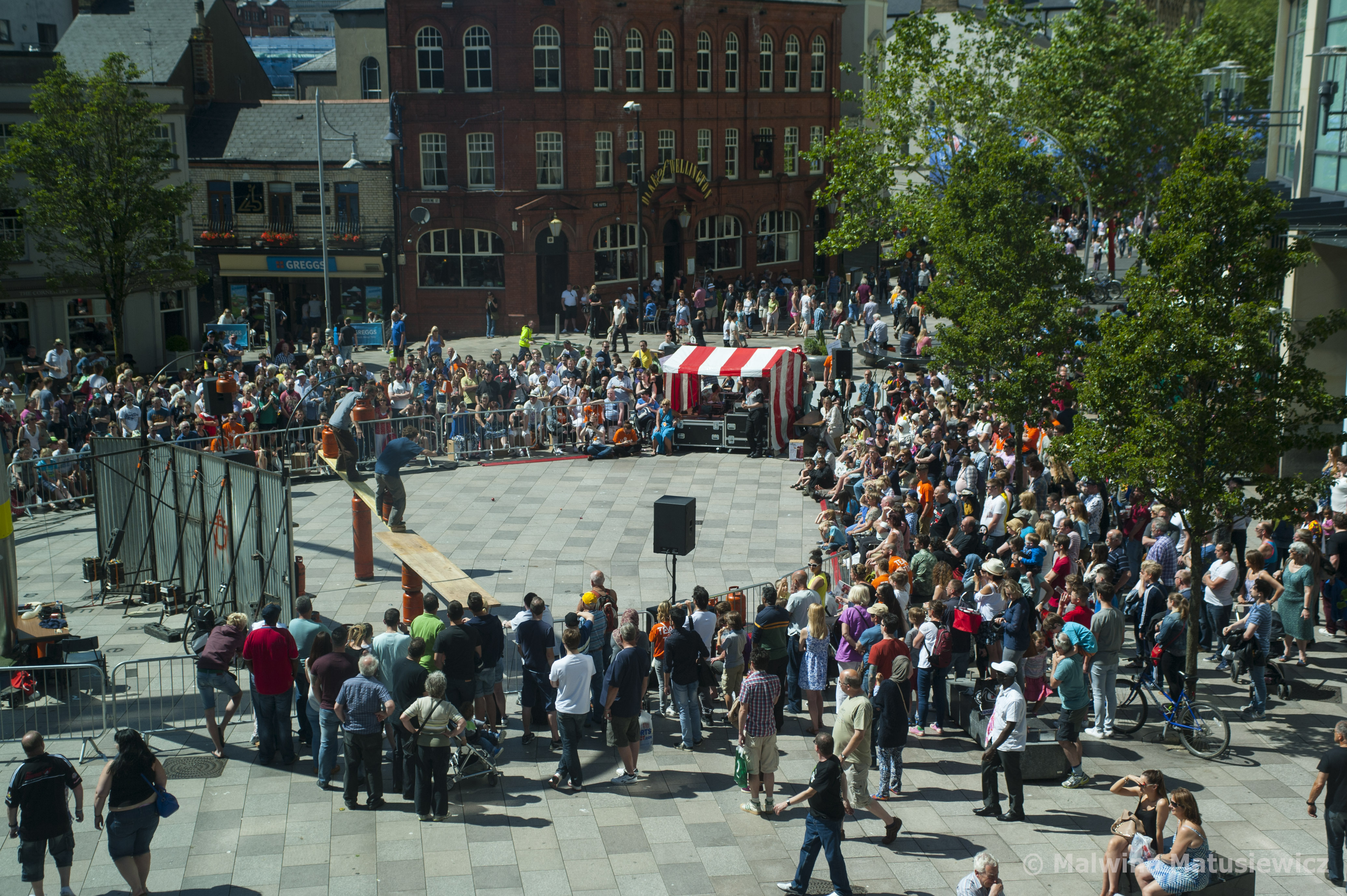 A group of people surround an act taking place in a town centre.