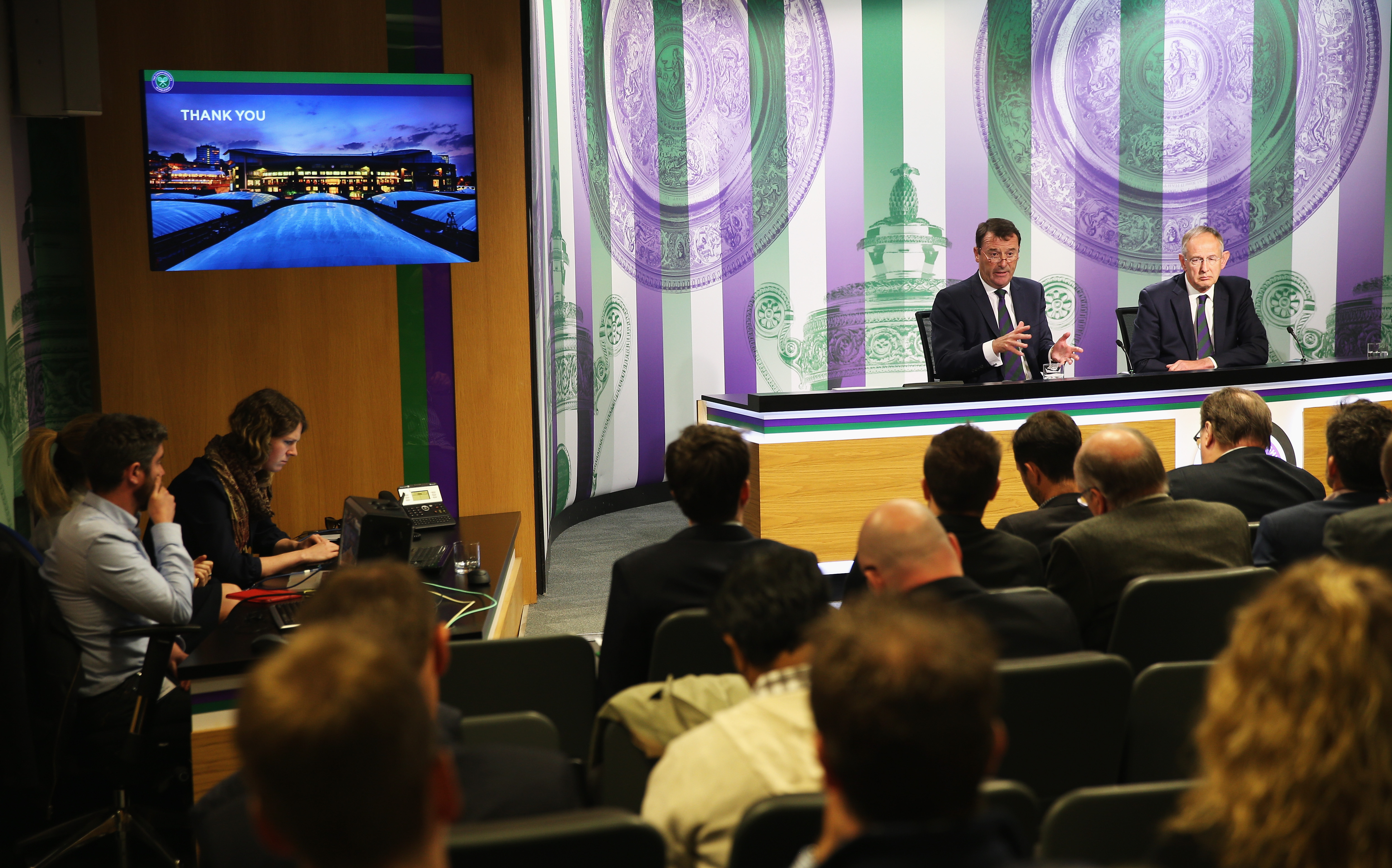 Media interview room with journalists questioning two people 
