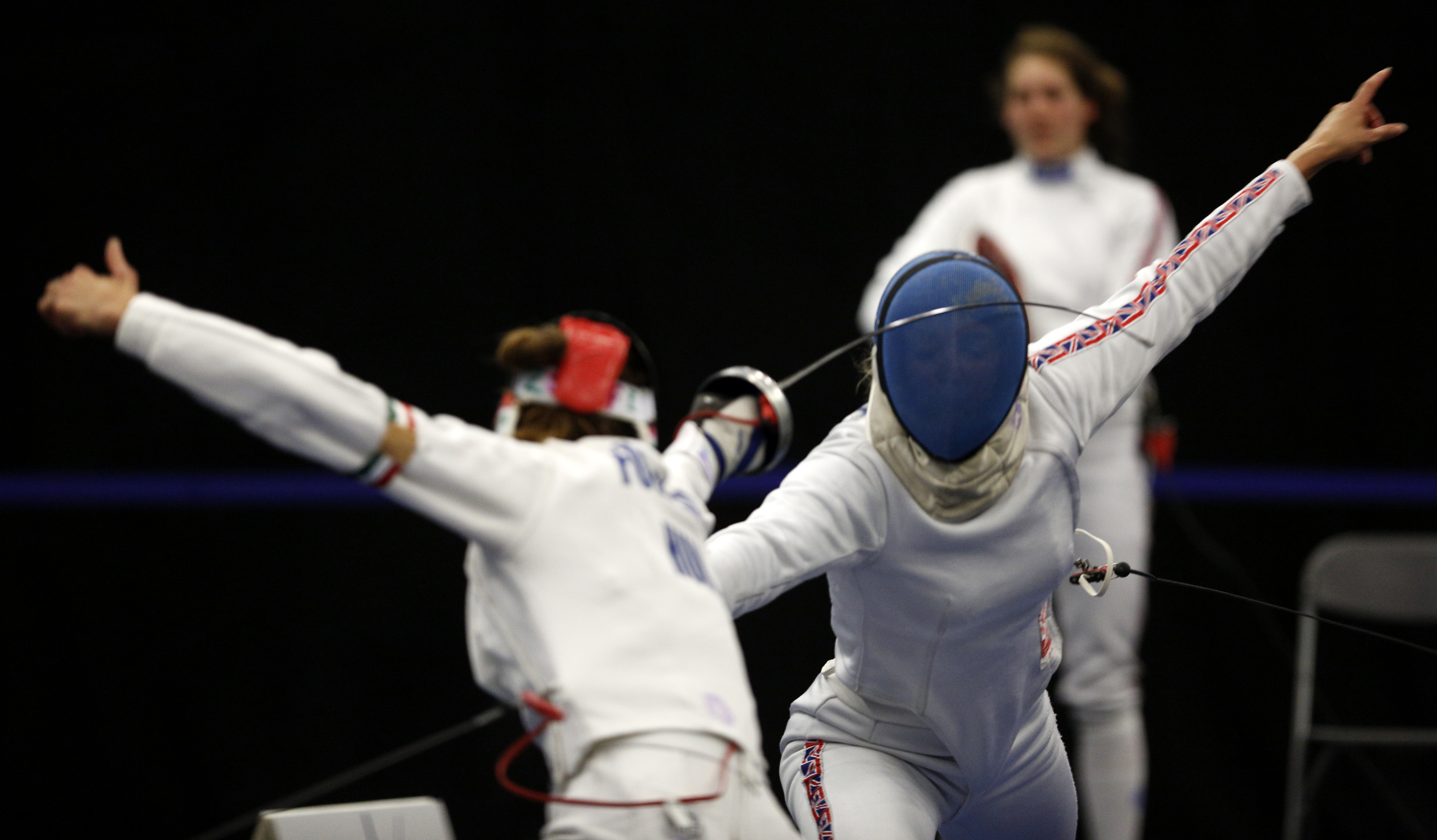 Two fencers lunge at each other in an arena, one face the camera with a blue mask.