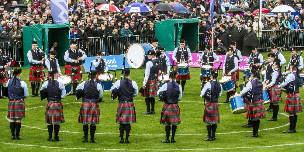 A band play in front of a crowd at a golf event, they wear kilts and traditional dress.