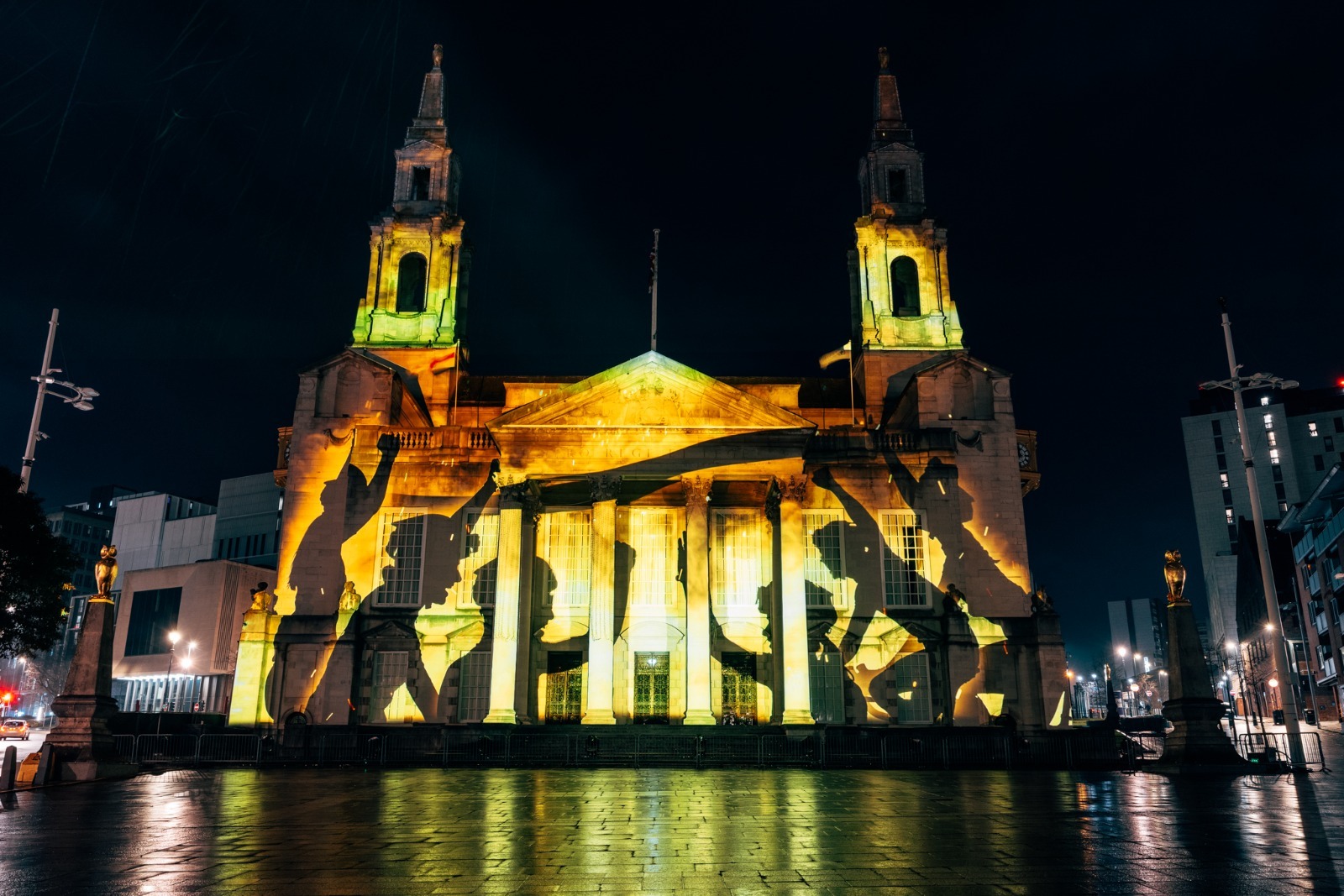 Leeds Civic Hall at night illuminated with a vibrant projection featuring silhouettes of cyclists in motion, set against a golden light display on the building’s façade. The wet pavement in the foreground reflects the lights, and the surrounding cityscape is dimly lit