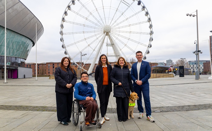 Group standing in front of a large Ferris wheel with a guide dog and a wheelchair user.